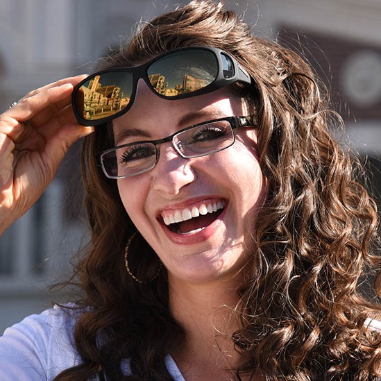 A woman wearing glasses smiles while raising her hands, highlighting a fashionable fitover that fits large prescription eyewear.